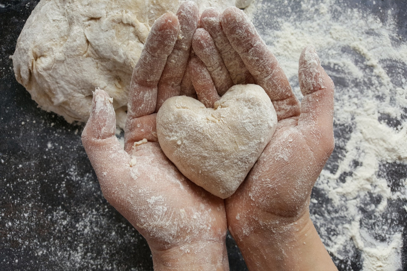 Dłonie obsypane mąką trzymające ciasto w kształcie serca na tle ciemnego blatu posypanego mąką, symbolizujące pasję do pieczenia w Cukierni Śnieżka w Gorzowie.