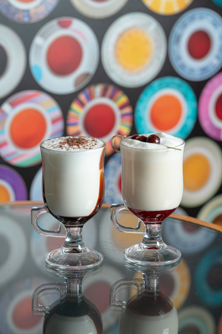 Dwa kubki z kawą i gorącą czekoladą, udekorowane bitą śmietaną i wiśniami, na tle kolorowych talerzy w Cukierni Śnieżka w Gorzowie.